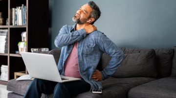 injured man sitting on the couch with his laptop grabbing his shoulder in pain