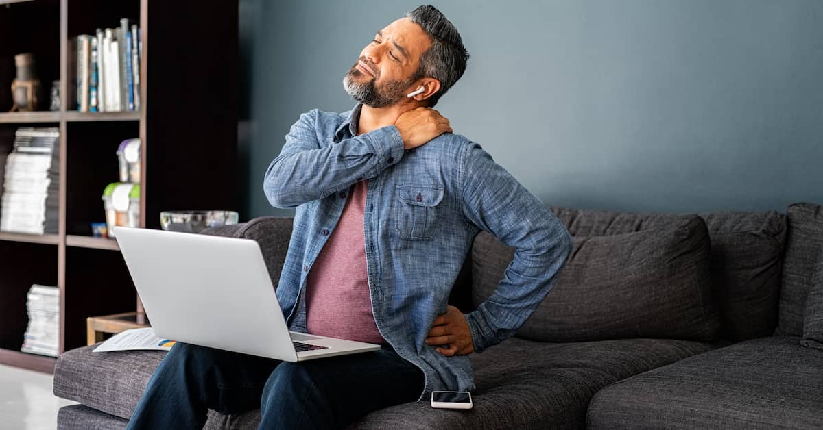 injured man sitting on the couch with his laptop grabbing his shoulder in pain